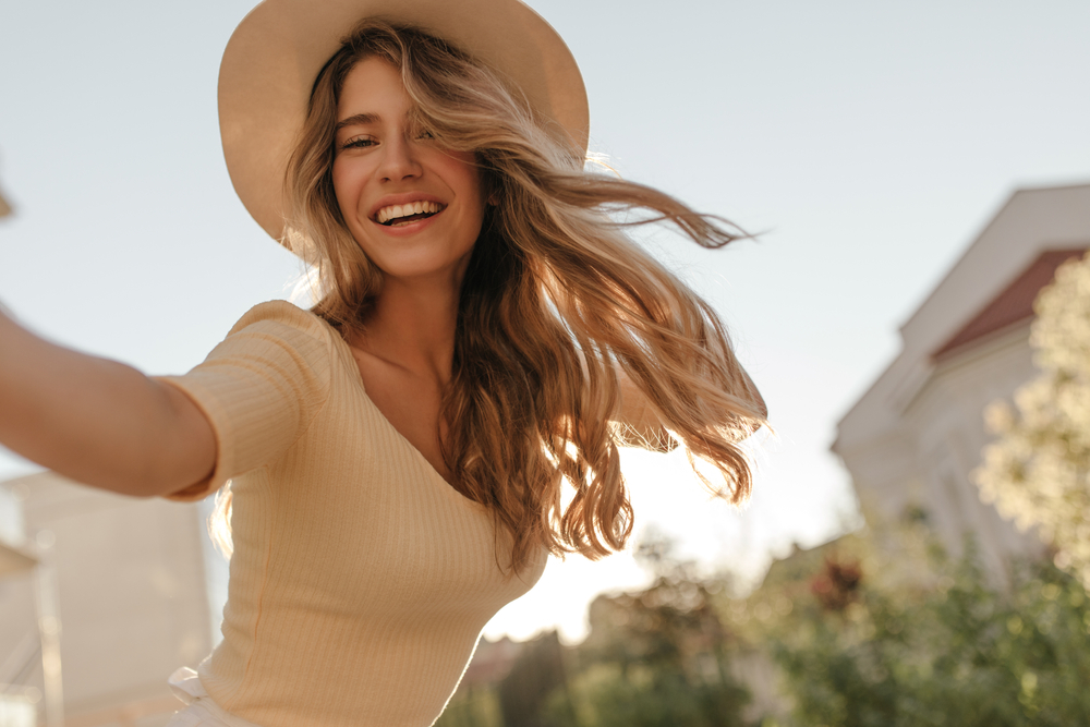 A cheerful woman with a wide smile and flowing hair, in a hat and yellow top, advertising Dental Veneers at Forest Ridge Dental Health & Wellness